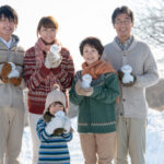 会社の福利厚生制度を利用して旅先で雪だるまを持ち微笑む3世代家族
